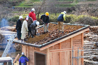 屋根の上に土や落ち葉を広げながら数人の作業員がスコップで作業している写真