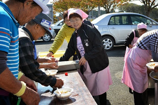 屋外のテーブルで、ピンクのエプロンをした女性たちが参加者に温かい豚汁を配っている様子の写真