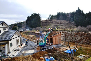 林に囲まれた開けた場所で、ユンボや複数の人が木造小屋の屋根建築の作業をしている写真
