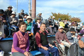 観客席で笑顔の女性と男性が座っていて、周囲にもたくさんの人々が腰かけて休憩している様子の写真