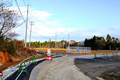 造成中の現場に電柱と仮設のガードレールが立ち並び、奥には整地された区画と資材置場らしきフェンスが見える造成地の進捗写真