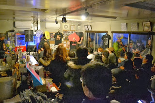 多くの観客が集まり、屋内で音楽ライブが行われており、ステージ上でギターを演奏する男性と桃の被り物をつけて歌を歌っている様子の写真