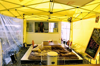 風よけに囲まれたテントの奥に厨房があり、井尻さんが顔をのぞかせていて、手前にはこたつが設置されているテント内の様子の写真