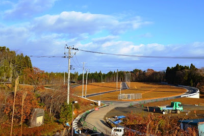 舗装された曲がり道に沿ってフェンスと資材が置かれ、奥に広がる区画整備された宅地が見える高台住宅地造成の写真
