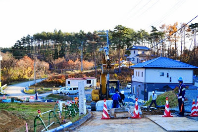舗装工事中の道路で作業員たちが重機の周囲で作業をしており、周囲には新築の住宅や電柱、カーブした道路があり、後方には紅葉した木々が広がる高台住宅地の工事中の写真