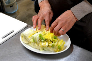 エプロンをした手が白い皿の上にある白菜の漬物を整えている様子が写っている準備中の写真