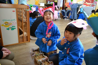 それぞれ手作りの鬼のお面をかぶった青いスモックの園児2人が一人は食べ物を口に入れようとし、もう一人は笑顔でカメラを見ている写真