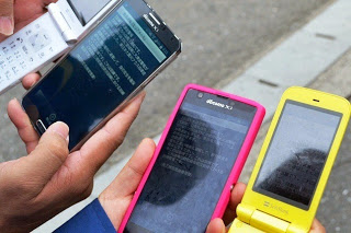 4人の手により、それぞれ異なる色や形の携帯電話やスマートフォンが緊急速報メールの画面を表示した状態で並べられている様子を写した写真