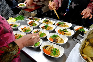 長方形の白い皿の上に大根や人参などの煮物を盛りつけている女性たちの手元が写っている盛り付け作業の写真