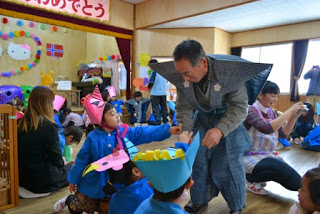 和装に大きな肩当てがある肩衣の衣装を着た男性が子どもたちに豆やお菓子などを配っている園内を写した写真