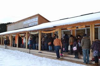 雪が積もる木造のポータルセンターの建物の前に多くの人が並び、イベント会場の受付を待っている様子の写真