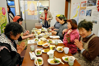 テーブルを囲んで数人の女性たちが笑顔で「いただきます」と手を合わせ、先ほど調理していた料理を前にして食事を始めようとしている様子の写真