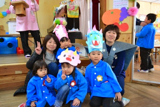 手作りの鬼の面をかぶり青いスモックを着た子どもたちが前列に並び、カメラに向かってピースサインをし保護者や先生が一緒に写り記念撮影をしている写真