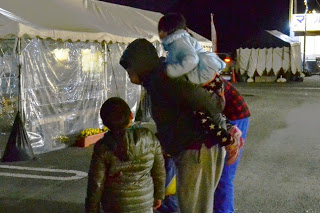 テントの前で、子どもを背負った男性と、女の子が並んで夜の会場を見つめている写真