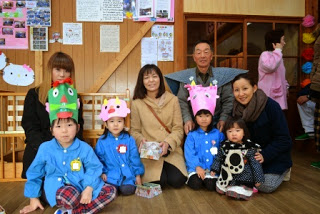 手作りの鬼の面の帽子をかぶった青いスモックを着た子どもたちと保護者や園の関係者と一緒に笑顔で一緒に記念撮影をしている写真