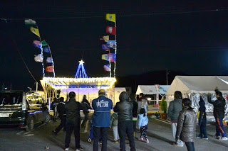 四隅に旗が設置され点灯されたステージの前に人が集まり、イルミネーションを見て楽しんでいる様子の写真