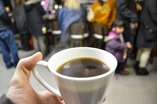 湯気の立ちのぼる温かいコーヒーが入った白いマグカップを手にもって見せている写真