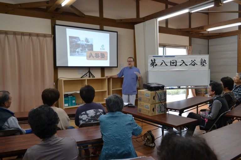 数人の高齢者が机に座って前方を見ており、スクリーンに映像が映し出され、『第八回入谷塾』と書かれたホワイトボードがある室内を写した写真