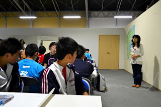 教室の前に立って話す女性の方を、生徒たちが椅子に座って静かに聞いている様子が写っている写真