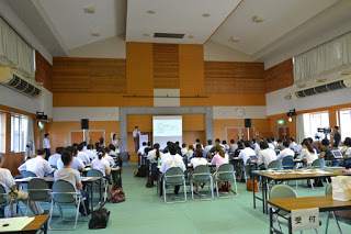 奥の壁に設置したスクリーンに資料が映し出され、大勢の参加者たちがスクリーンの方を向いて座っている会場全体の様子を写した写真