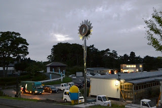 夕方の曇り空の下、中央にひまわり型の時計台が立ち、駐車場には多くの車が停まり、奥には森に囲まれた建物が点在する施設周辺の風景の写真
