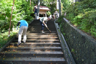 長くて急な石段を、2人の方がほうきで掃除している様子で、階段の先には神社の拝殿が見える写真