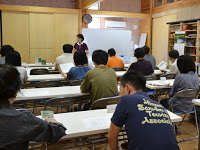 教室で複数の生徒たちが机に座って前方のホワイトボードの前に立つ教師の話を聞いている様子の写真