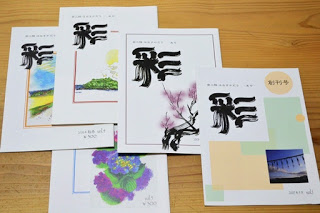 木の机の上に、それぞれ異なるイラストや写真がデザインされている「彩」という文字が表紙に大きく書かれた冊子が5冊重ねて並べられている写真