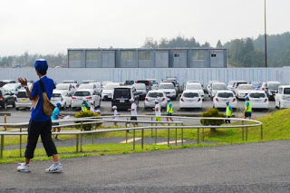 広い駐車場の前を、白い体操服を着た子どもたちが引率者とともに列になって走り抜け、手前では青い服のスタッフが拍手で応援している1000キロメートル縦断リレーの写真