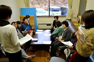 長机に資料が置かれ7名の女性がソファーに座り、2名の担当の女性が説明をしている写真