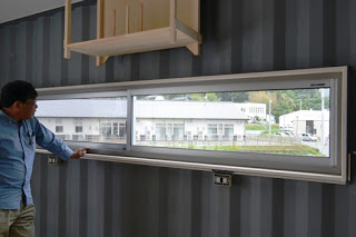 室内の壁に設けられた横長の窓のそばに鈴木清美さんが立っており、窓の外には建物や緑が見え、窓の上には木製の棚が取り付けられている写真