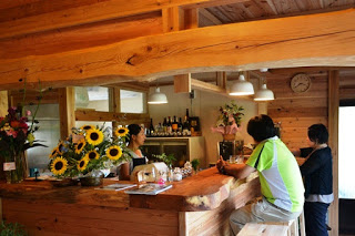 中に女性が立っている、ヒマワリの花が飾られたL字型の木のカウンターに一人の男性が座り、奥に女性が立っている写真