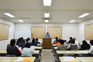 明るい教室で中央に立つ年配の男澤達夫さんが話をしており、机に座った数十人の参加者が真剣に耳を傾けている写真