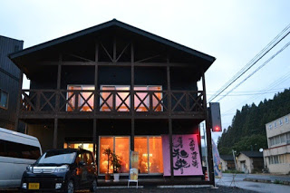 夕暮れの中、前に車が停まった木造の二階建ての建物の大きな窓から明かりが漏れ、右側にはピンク色の大きな布看板設置されている写真