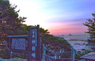 緑豊かな木々に囲まれた場所に説明版や看板が設置され、朝焼けに染まる海岸が静かに佇む様子を写した神割崎の写真