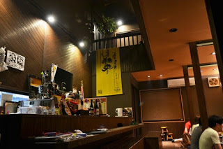 居酒屋の店内の酒瓶などが並んだ木製カウンターとその前に並ぶ客席が写っており、温かみのある照明が空間を照らしている様子の写真