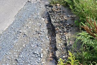 アスファルトと砂利が混ざった道の端に、雨水などで削られてできた溝があり、そこに小石や水がたまっている様子の写真