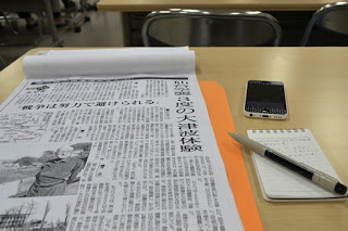 机の上には仙台空襲 3度の津波体験の見出しと男性の写真が載っている新聞記事とメモ帳、ボールペン、携帯電話が置かれている写真