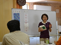 ホワイトボードの前に立って本を持っている女性と、席に座って女性の方を見ている男性が写っている写真