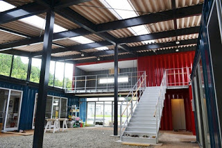 コンテナを組み合わせて中庭型に配置した建物の内部で、中央に白い階段があり、吹き抜けの天井から自然光が差し込む内観の写真