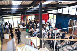 コンテナを組み合わせた建物の広々とした中央スペースに多くの関係者の方々が椅子に座って集まっている開所式の様子の写真