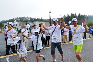 道路わきで応援している人たちの前を、色とりどりのタスキをかけた5名の大人と子どもが楽しそうにつないだ両手を上げて走っている写真