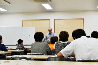 教室内で男澤達夫さんが正面の教壇に立って話しており、手前には子どもから大人まで複数の参加者が座って真剣に耳を傾けている様子が写っている講演中の写真
