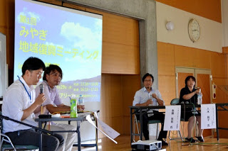 資料が映し出された中央のスクリーンを挟んで4人の男女が壇上の席に座っており、一番右側の男性がマイクを持ち、パネルディスカッションを行っている写真