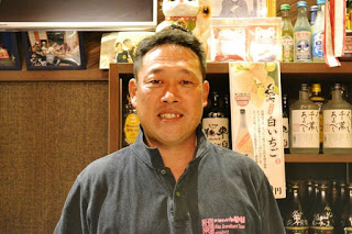 たくさんの酒瓶が並んだ棚の前に笑顔で立つ佐々木さんの写真