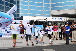 色とりどりのタスキをかけた5名の大人と子どもがつないだ両手を高く上げてゴールした瞬間を後方から写した写真