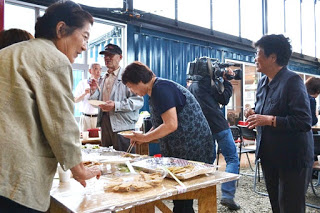 コンテナ建築の屋内スペースで数人の高齢女性が料理を囲みながら楽しそうに会話をしており、背景にはカメラを構えて撮影している人も見える写真