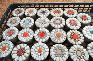 黒いプラスチック製のトレイの中に複数の円形コンクリート製敷石が並べられており、それぞれの敷石には白い小石で花の模様が描かれ、中央には赤・青・緑・オレンジ・茶色などの色付きの石やタイルが配置されているエコ平板の写真