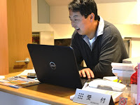 受付カウンターの中で、黒いノートパソコンの画面を見ながら笑顔で作業している佐々木さんの写真