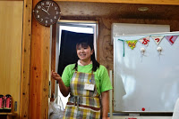 ホワイトボードの前に立っている黄緑色のTシャツとチェックのエプロンを着た女性が、笑顔で話している様子の写真